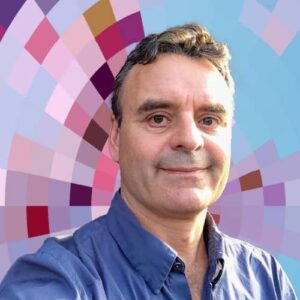 Headshot of Nate Angell, a smiling white man with brown hair wearing a blue shirt on top of a MYFest background of colorful pink, purple and blue four-sided shapes spiraling out from the center.