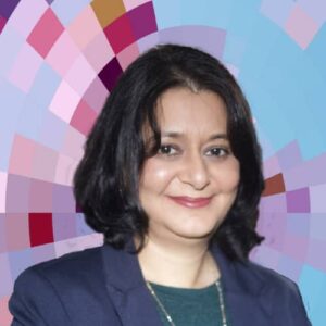 Headshot of Sukaina Walji, a smiling woman with medium-length, dark hair wearing a blue suit jacket over a green shirt and a silver necklace, on top of a MYFest background of colorful pink, purple and blue four-sided shapes spiraling out from the center.