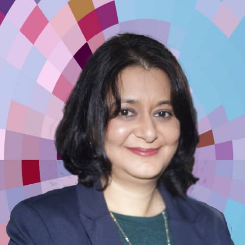 Headshot of Sukaina Walji, a smiling woman with medium-length, dark hair wearing a blue suit jacket over a green shirt and a silver necklace, on top of a MYFest background of colorful pink, purple and blue four-sided shapes spiraling out from the center.
