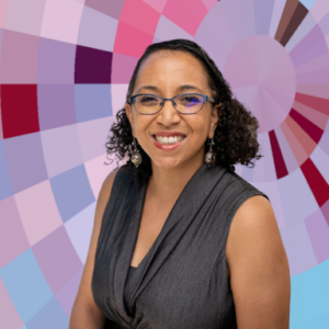 A woman with dark, shoulder-length curly hair and brown skin. She is smiling showing teeth and wearing blue-rimmed oval glasses, silver spherical earrings, and a grey dress.