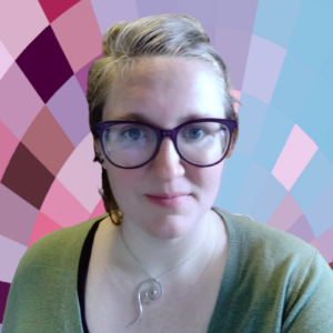 A white woman with her hair (a mix of natural reddish brown with a significant sprinkling of white-grey) tied back, large purple-framed glasses, and an awkward smile. She is wearing a black shirt with a light green cardigan on top and a Goddess Spiral necklace.