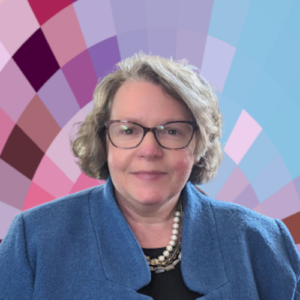 Sarah is a white woman with medium-length light brown hair. She is wearing a black top, a blue jacket, with a pearl and gold necklace. She is wearing glasses.