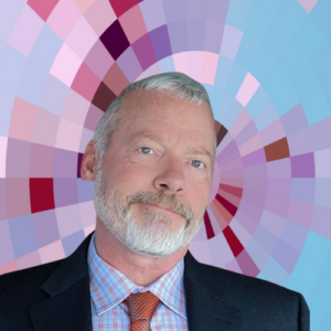 Headshot of Shaun Longstreet, a smiling man with short white hair, beard, and moustache wearing a dark suit, patterned shirt and red tie on top of a MYFest background of colorful pink, purple and blue four-sided shapes spiraling out from the center.